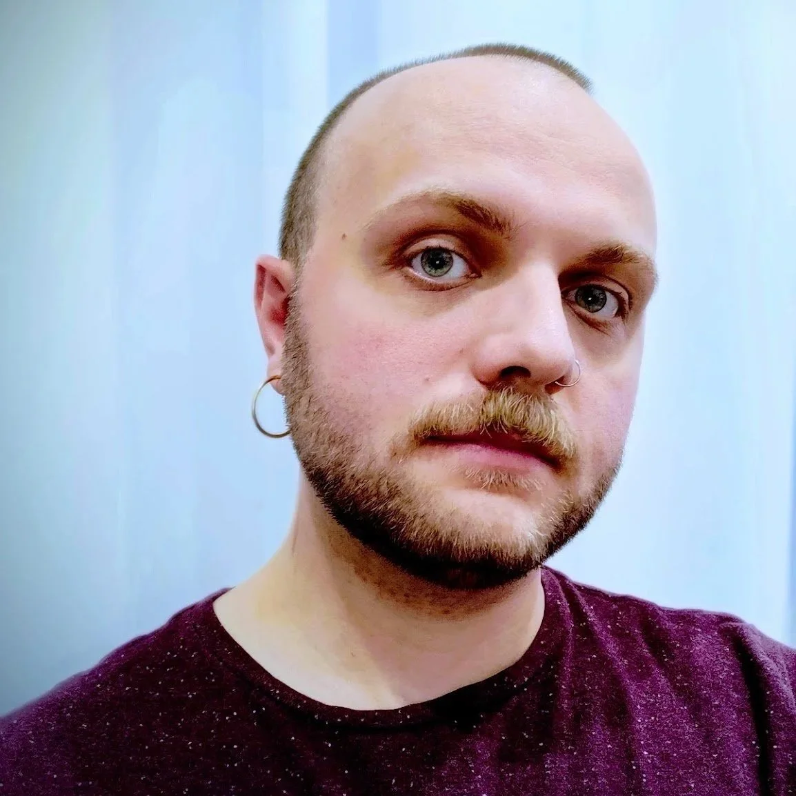 Photo of Jaspen, a nonbinary white person with short, buzzed, brown hair, mustache, and beard, wearing a hoop earring, and a burgundy-colored shirt. The background is a white curtain.