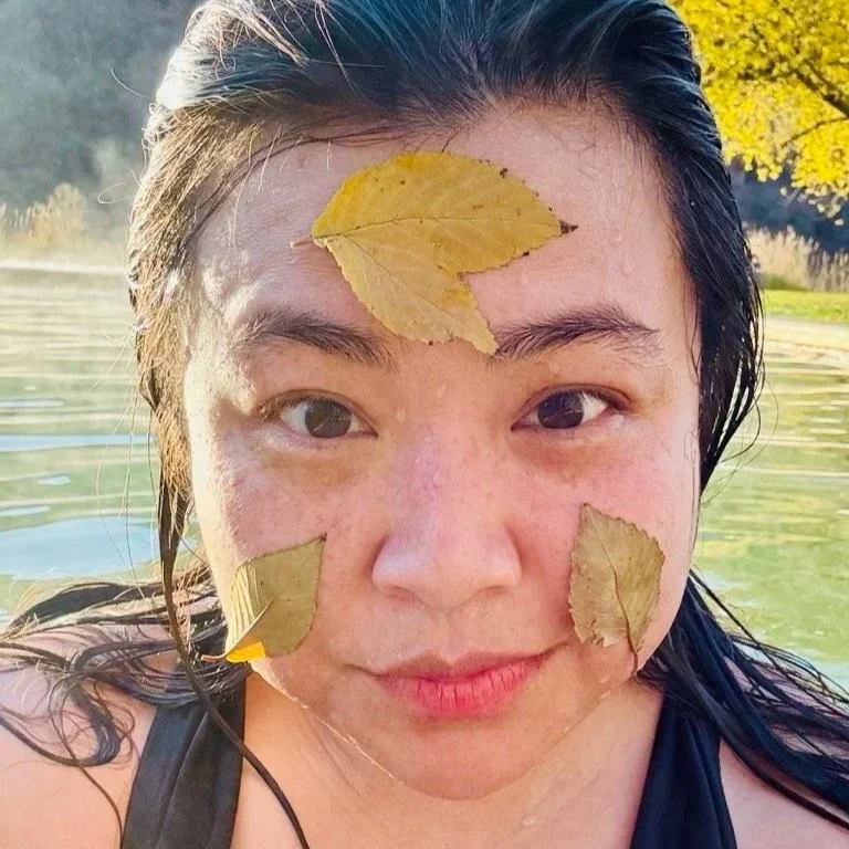 Sophia is wet and Asian. They have damp face and flattened hair. Yellow leaves are stuck to their cheeks and forehead. There is a green-tinted body of water behind them, nestled in a hilly landscape.