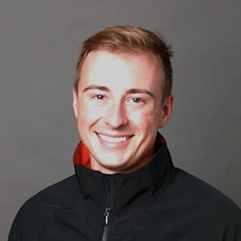 Nathan is a white, male-presenting person with short brown hair. He is smiling and wearing a black zip-up jacket with red inside the collar. They stand against a solid grey background.