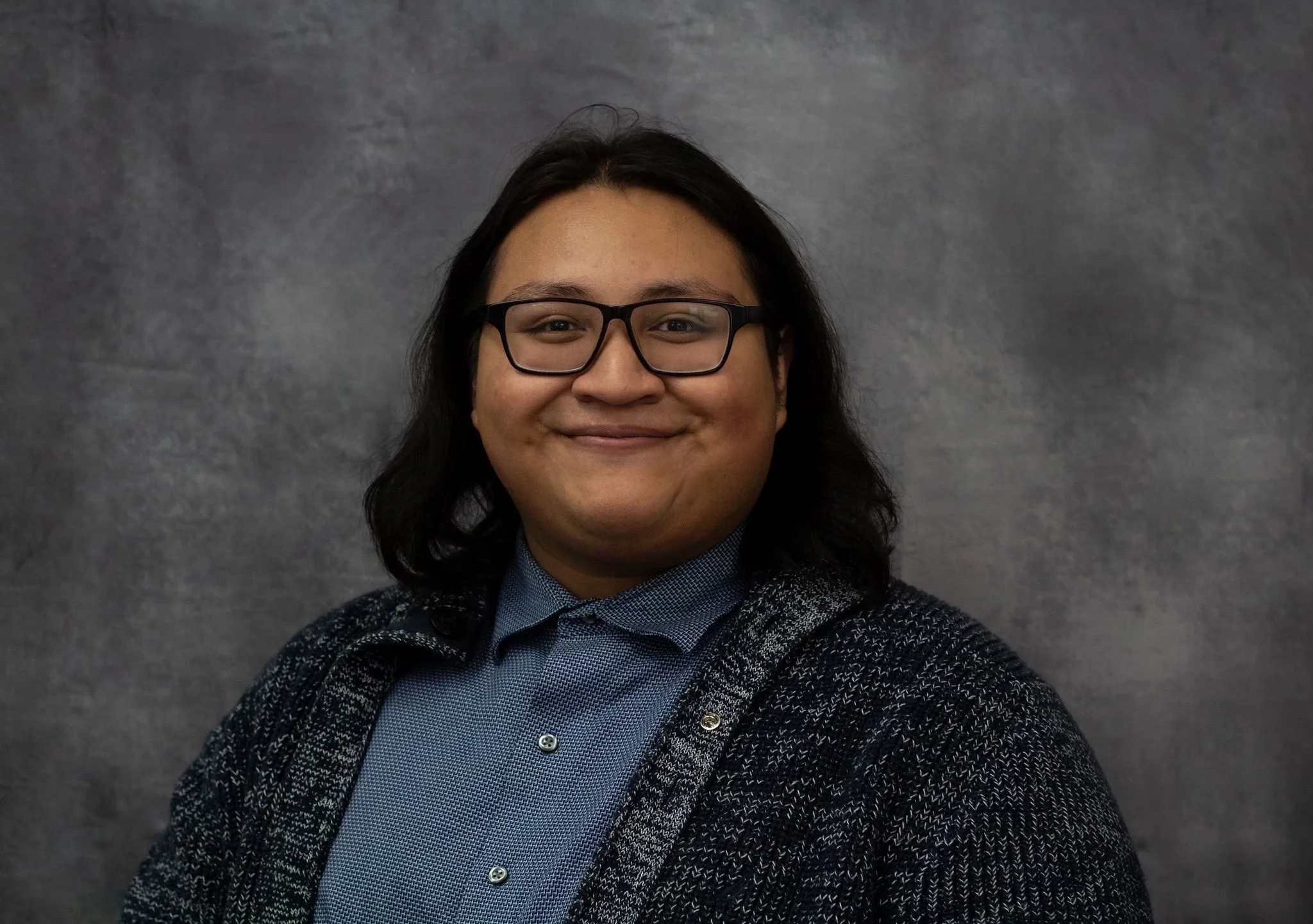 Pablo is a cis gender male with long, dark hair and black rectangular glasses. He wears a blue button-up shirt and dark textured cardigan and smiles gently at the camera against a softly blurred gray background.