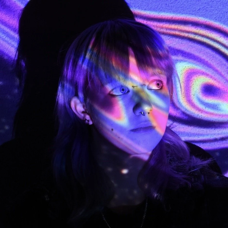 Raine is a gender queer white person with medium length blue hair and bangs. They're wearing a grey button up jacket, and looking to the right of the photo with a neutral expression. A rainbow overlay covers their face and the background.