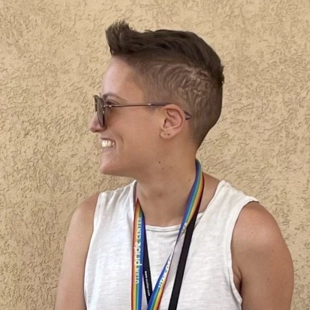 Ro is a genderqueer person with short brown hair. Ro's face is looking to the right and their left side profile is in the photo. They're wearing sunglasses, a white shirt, and a rainbow lanyard against a beige background.