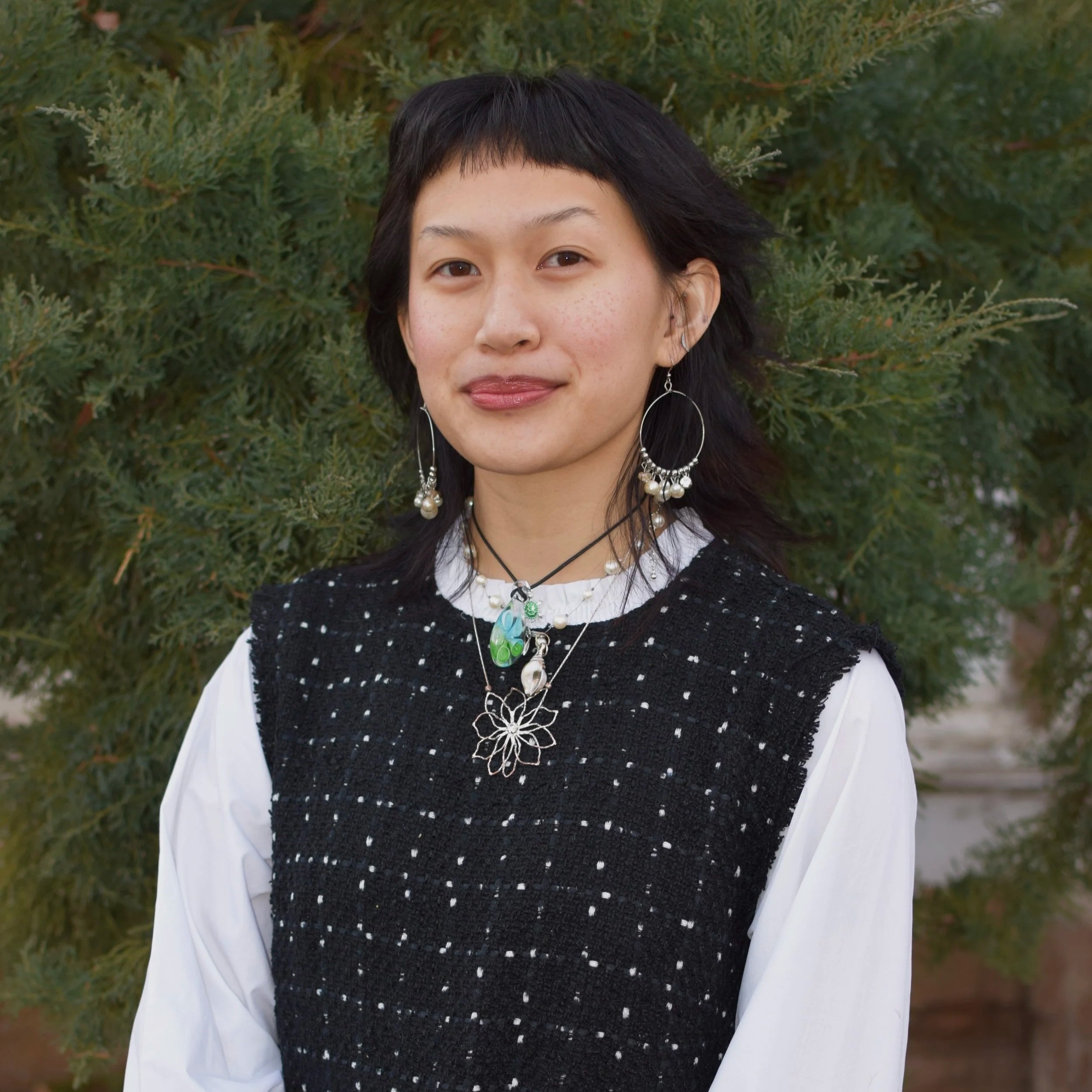 Zabrina is a Vietnamese-American woman with medium length black hair and micro-bangs. Zabrina is wearing a dress with white long sleeves and a black vest-like layer. She is placed in front of a juniper tree smiling softly.