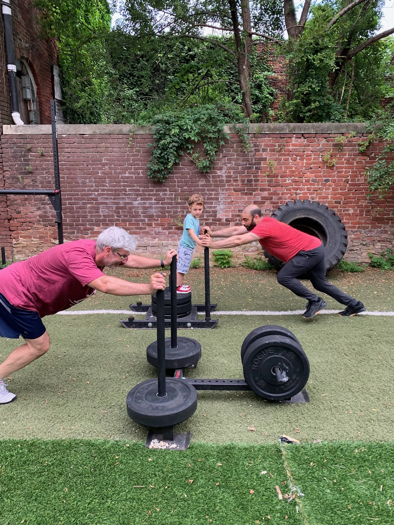 Two men and a boy working out with weightlifting equipment outdoors, pressing a small barbell while the boys stands on a weight sled, with a brick wall, large tire, and trees in the background.