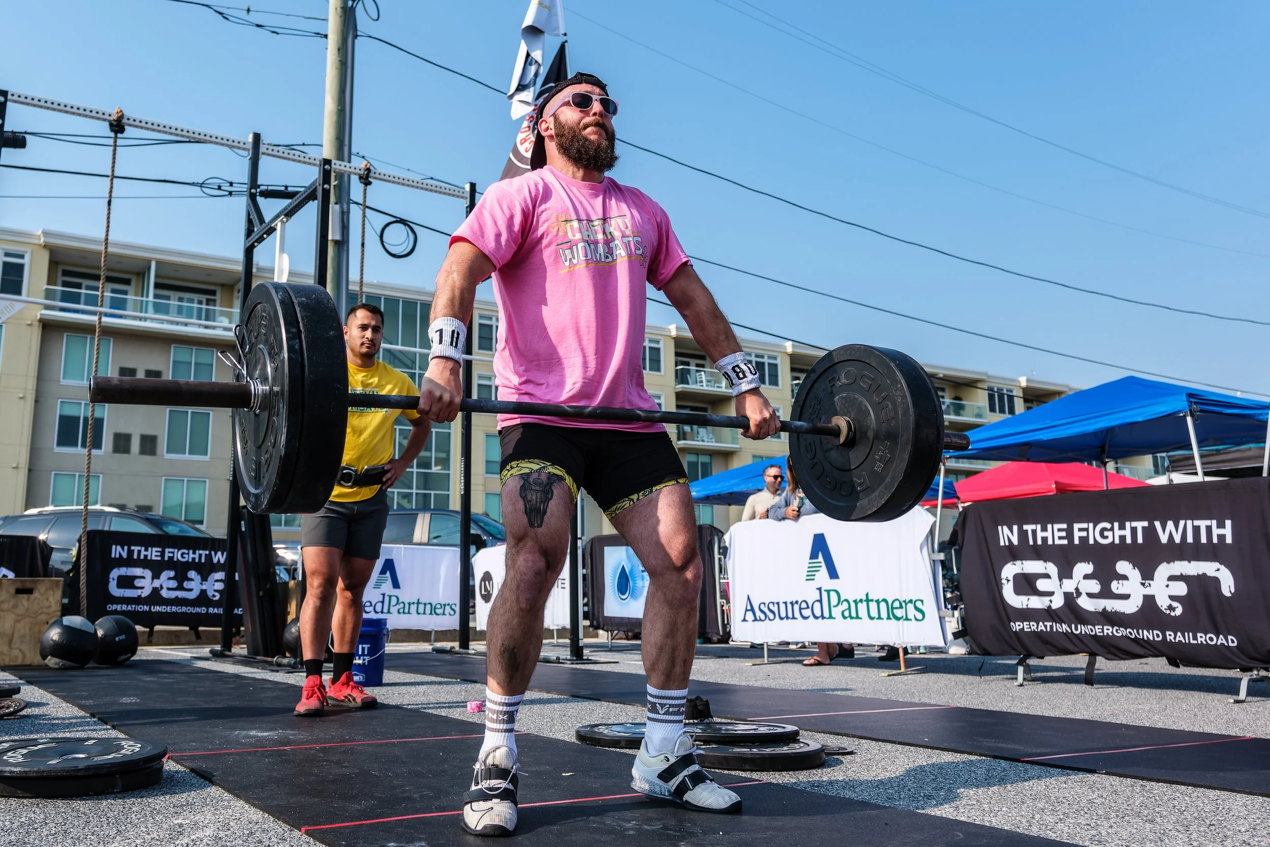 Man lifting a barbell during a strongman competition outdoors on a bright day, with spectators and banners in the background.