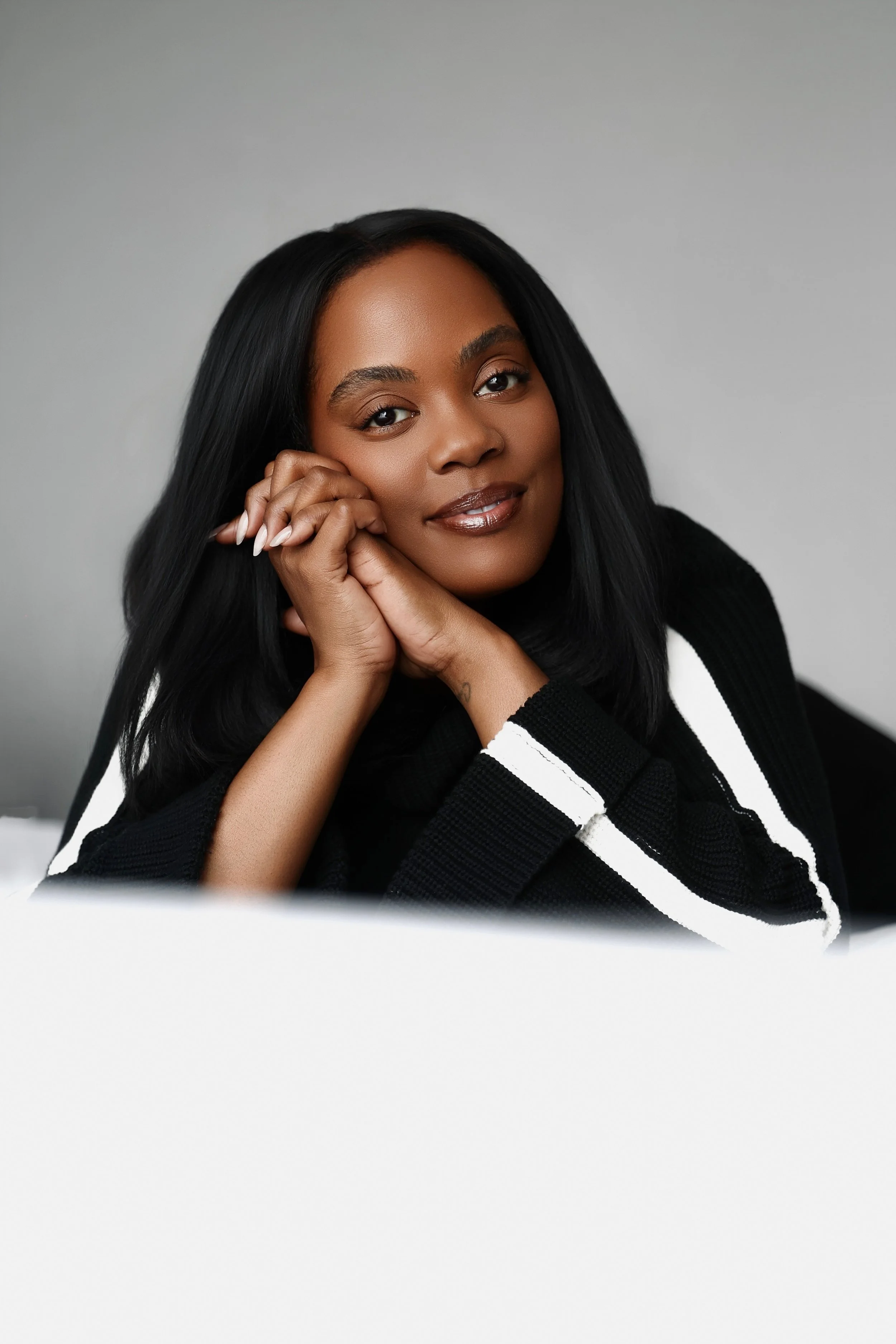 Close-up portrait of a Black woman with long black hair, smiling softly, resting her chin on her hand, wearing a black and white striped top, against a gray background.