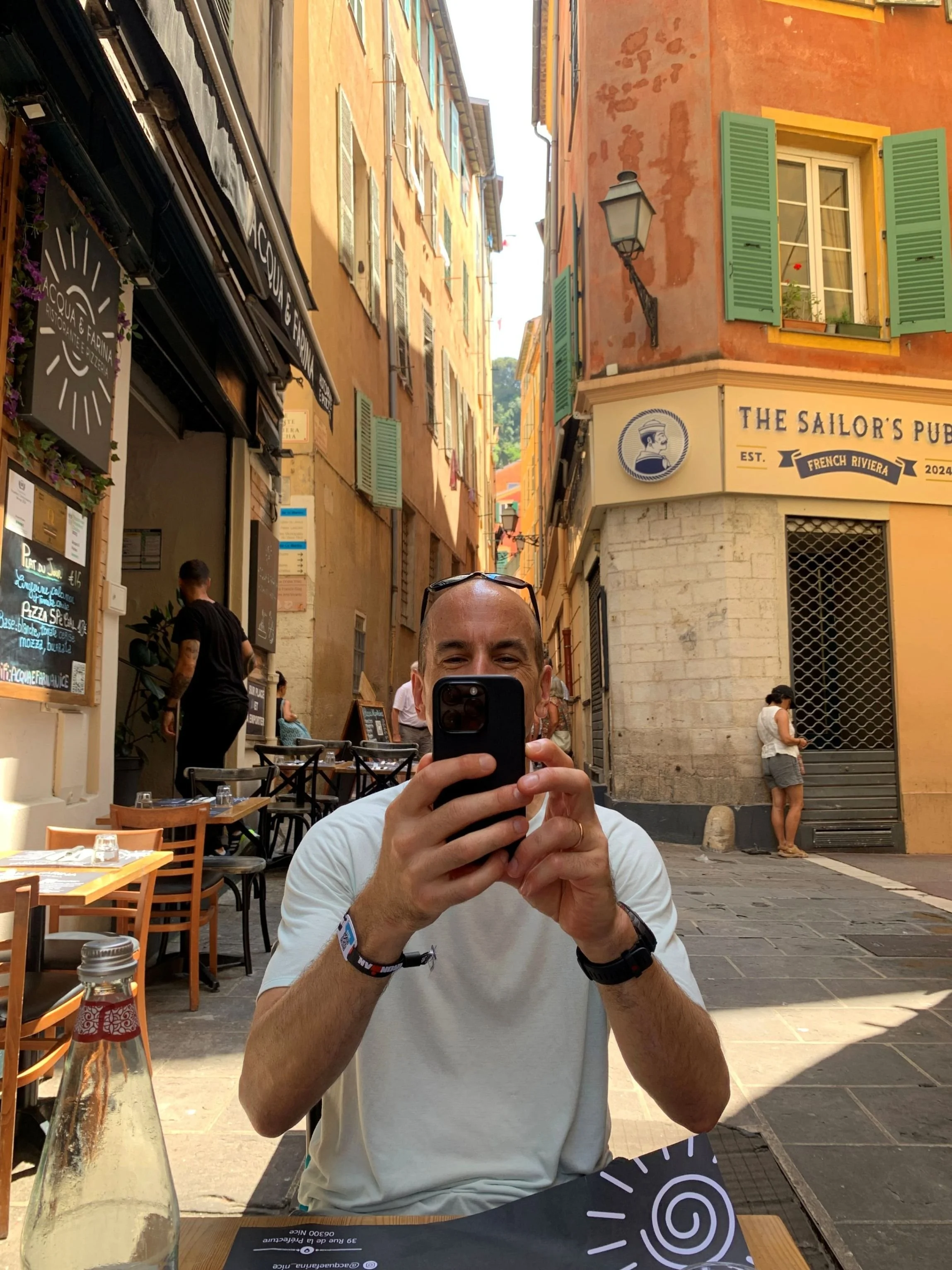 Man seated across a table in a pedestrian area holding a phone in two hands to take a photo of the person taking the photo.