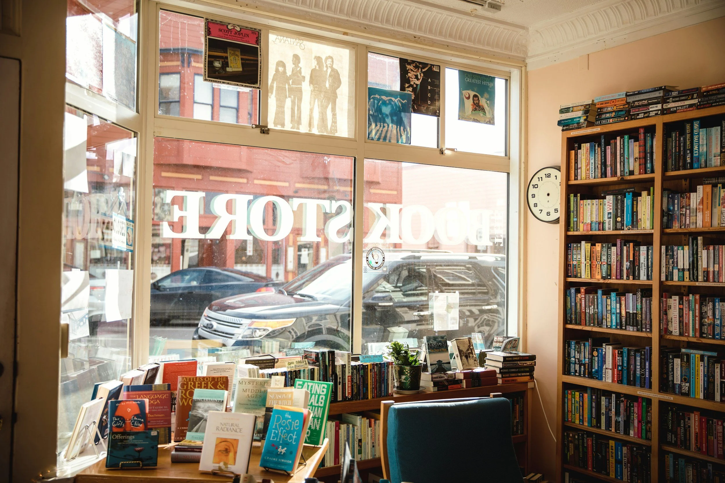 Image taken from within a bookstore, looking toward the front tables with books on display stands and out the window into an urban street beyond.