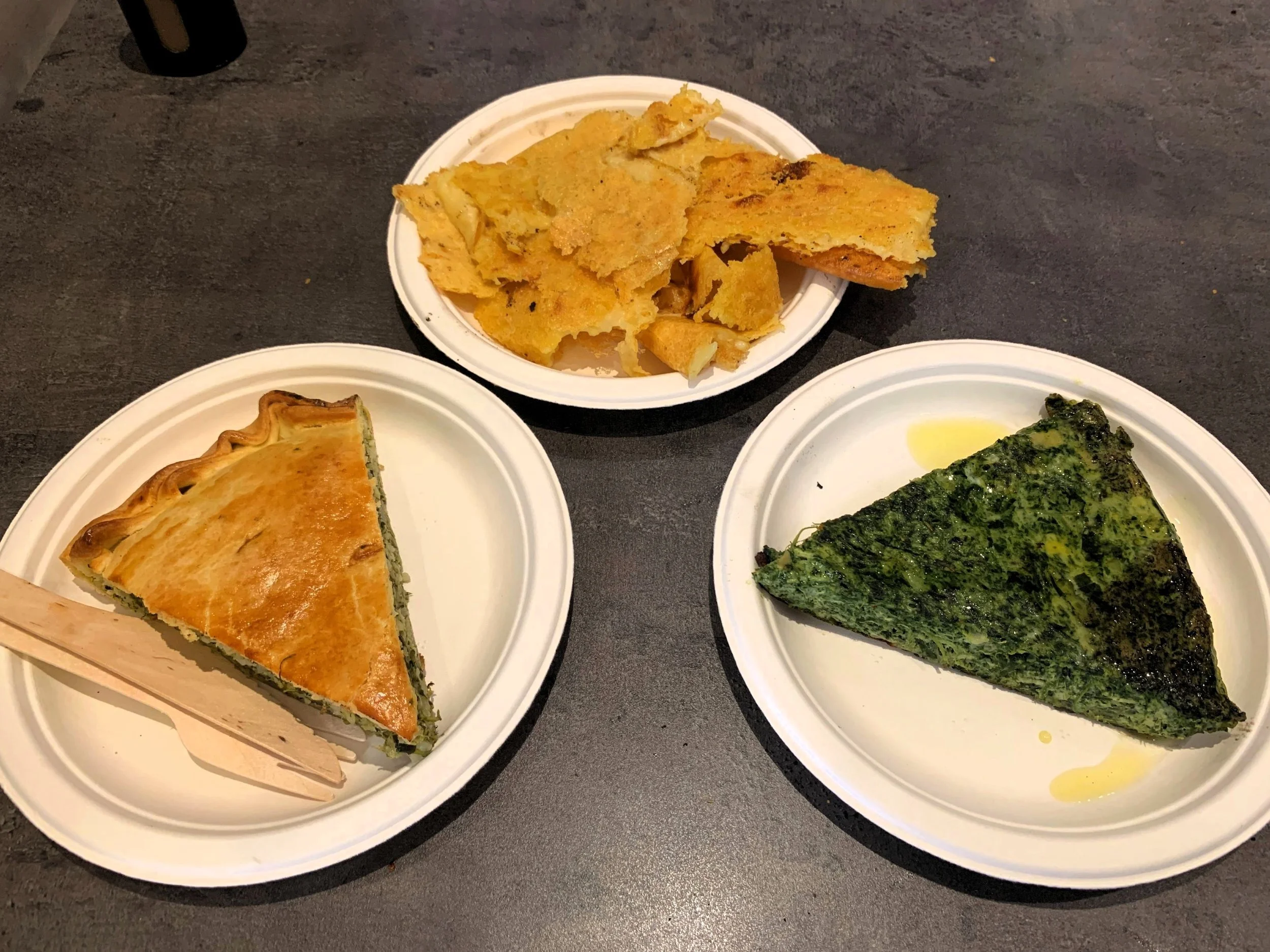 Three paper plates arranged in a triangle, two with triangular slices of pie-like foods and one with a hashwork of irregular thin slices.