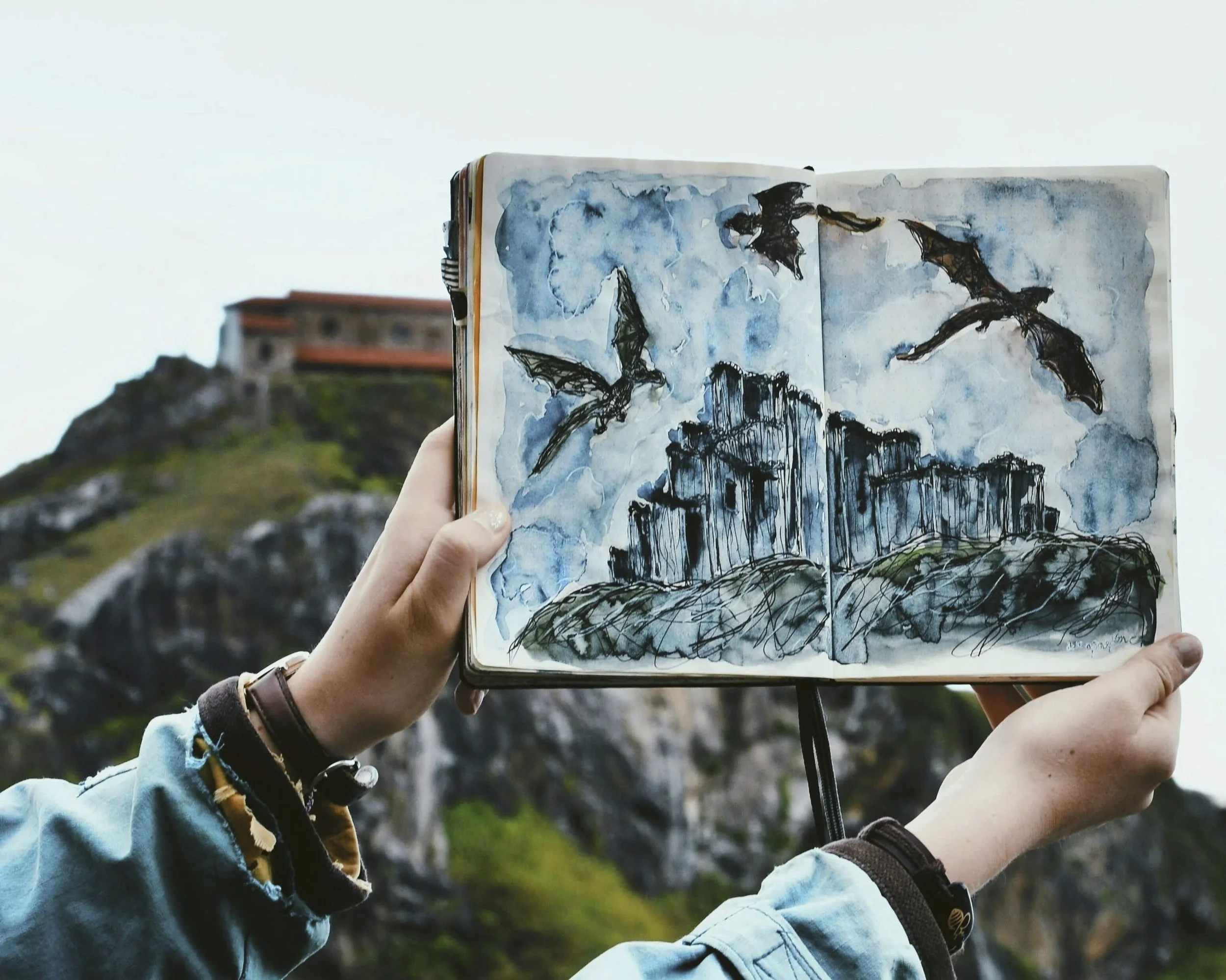Hands holding a sketch of dragons flying over a cliff with a cliff in the background.