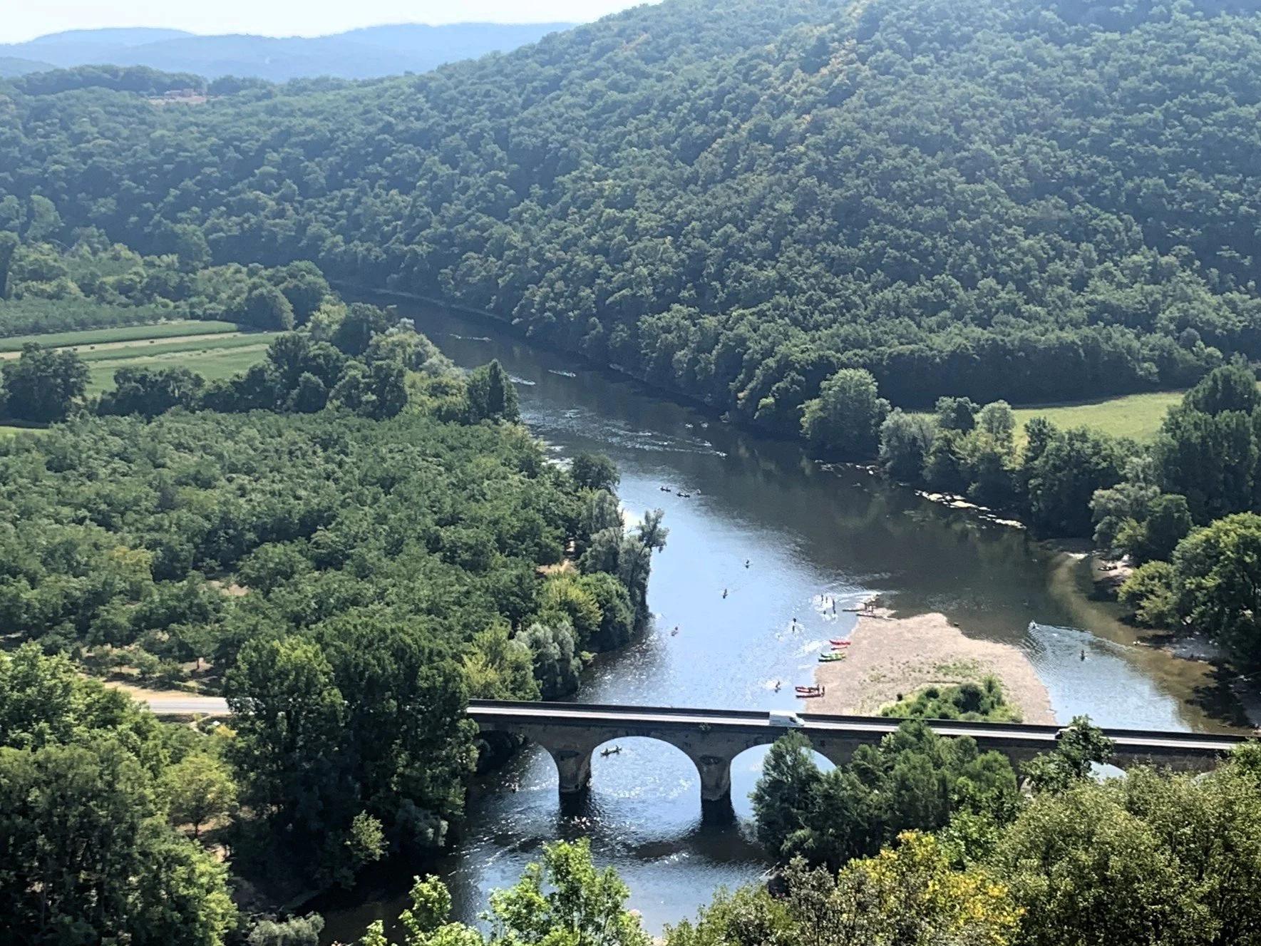 View from on high of a narrow bridge spanning a river dotted with canoes and kayaks, surrounded by fields and forest.