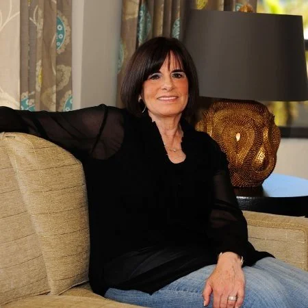 A woman with short dark hair sitting on a beige sofa in a living room. She is wearing a black top with sheer sleeves and jeans. There is a black lamp and a decorative gold vase behind her near a window with patterned curtains.