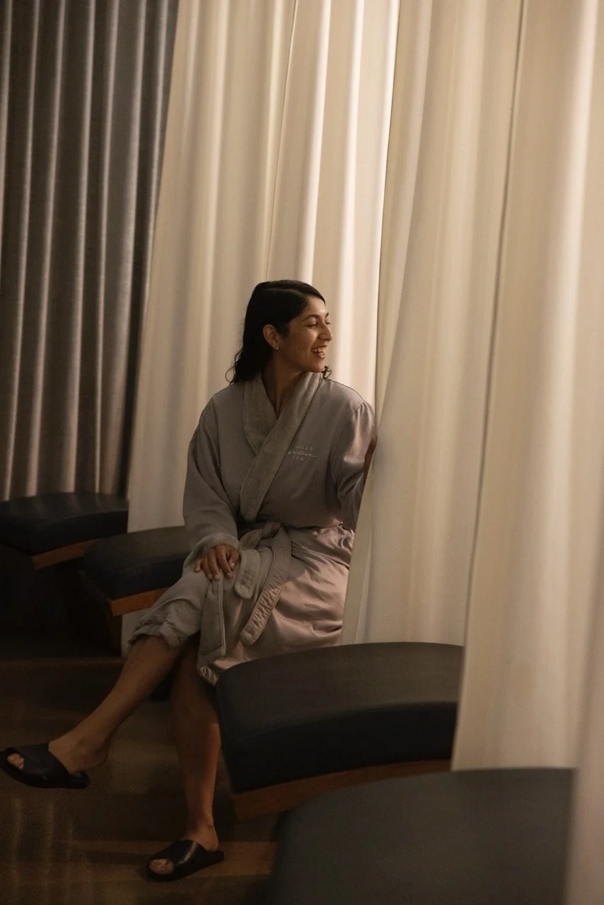 Woman sitting on a bench in a robe, smiling and looking to the side, next to beige and brown curtains.