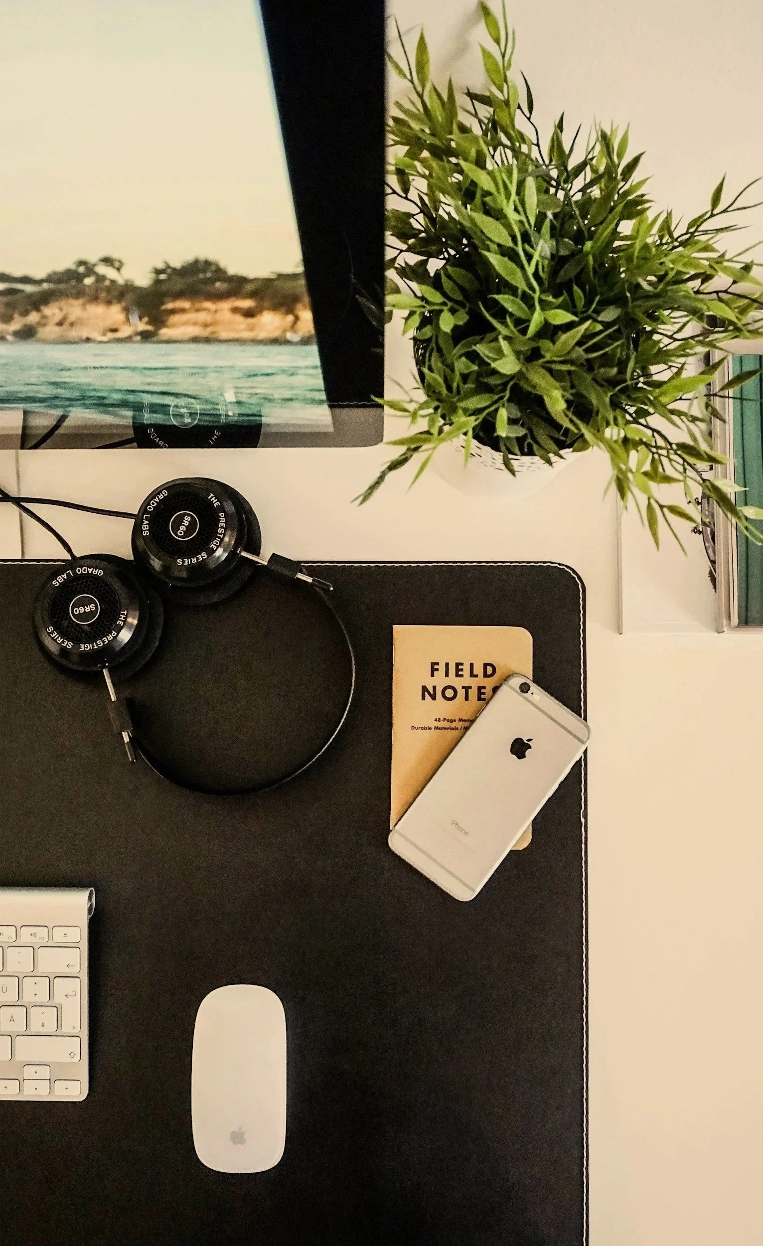 An overhead view of a clean workspace featuring a black desk mat, open-back headphones, a small brown Field Notes notebook, a silver smartphone, and a potted green plant in the corner.