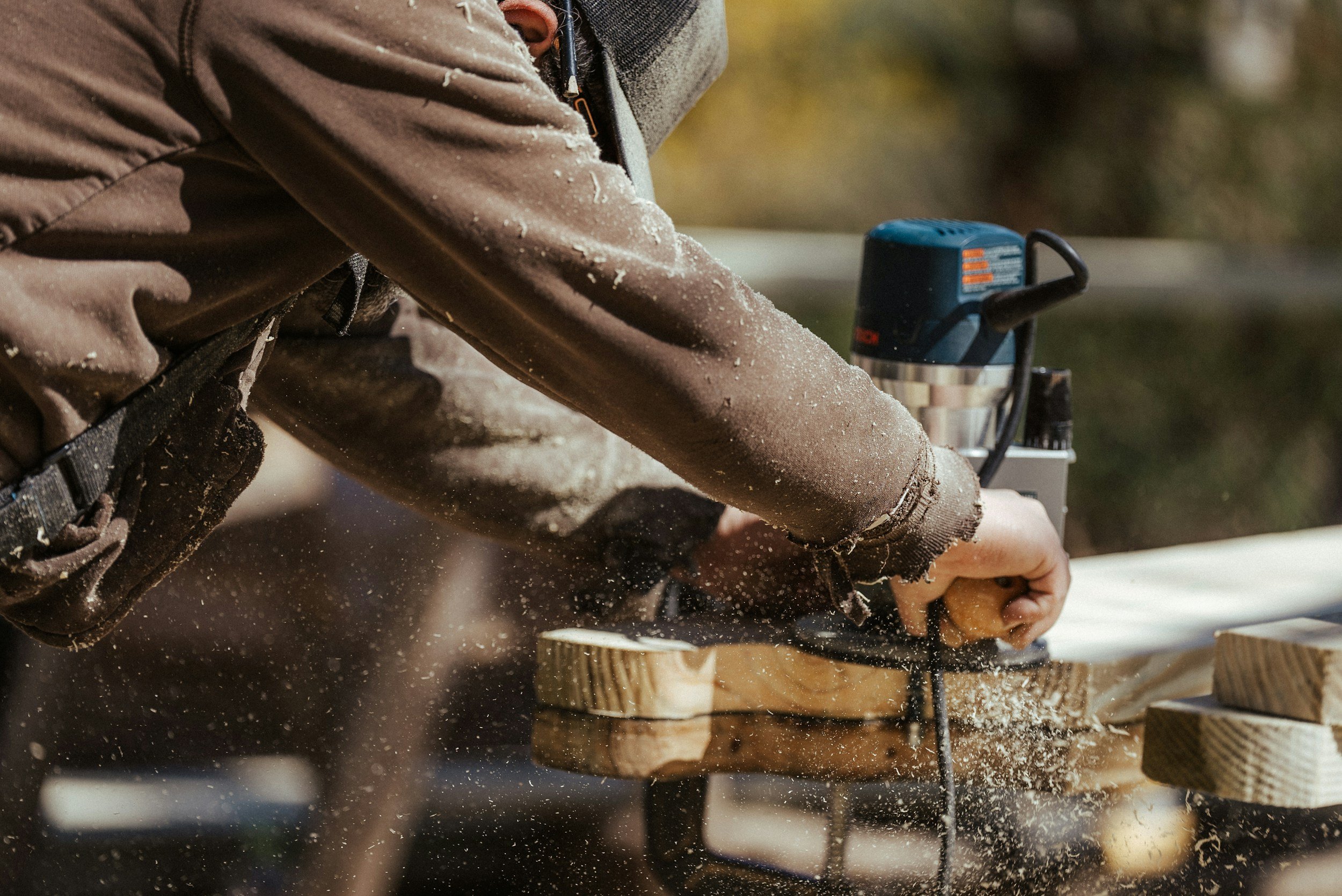 Close-up of a woodworker using a router on timber, with sawdust flying through the air and hands gripping the wooden handles for a precise cut.