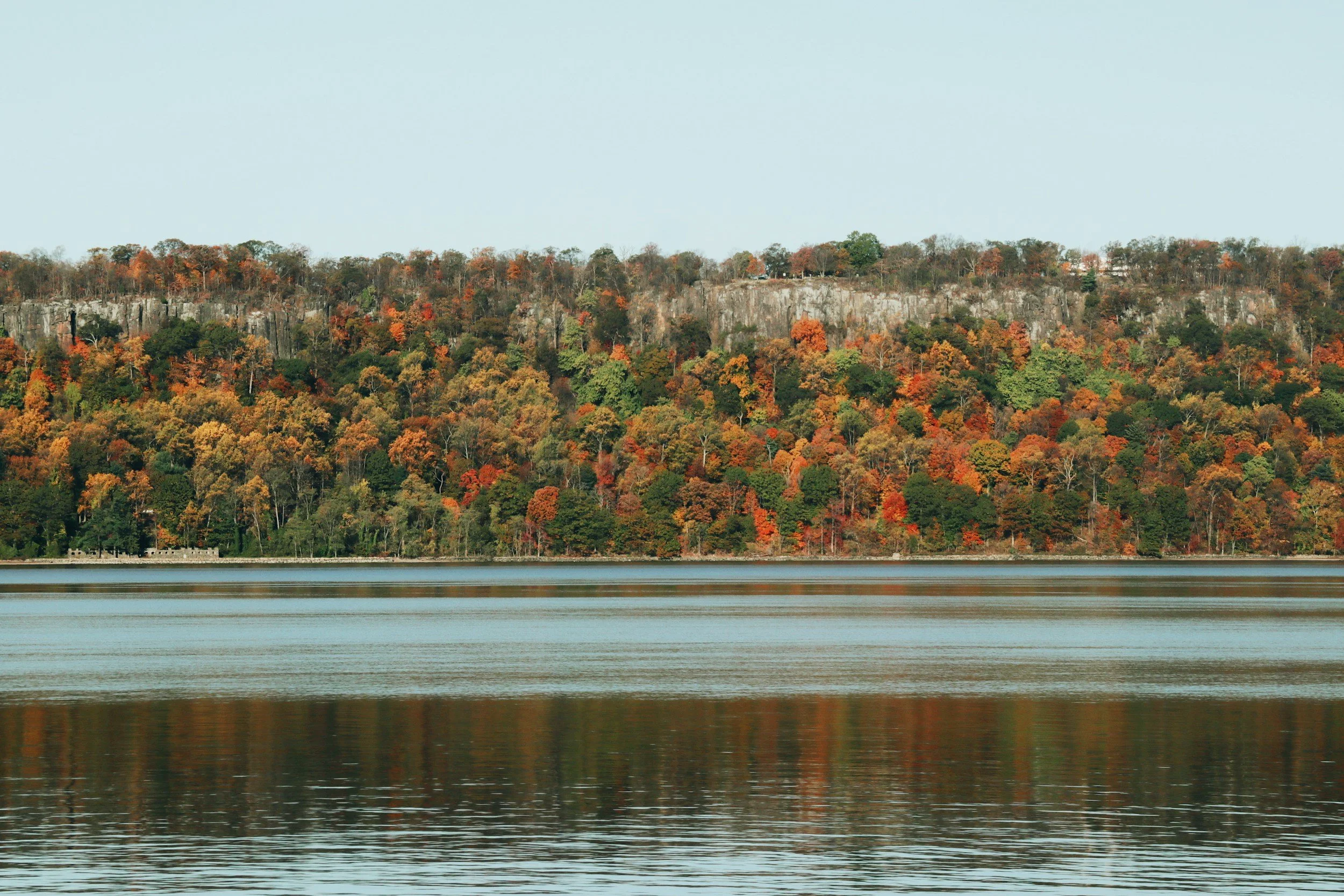 A panoramic view of the Hudson Valley in autumn, featuring vibrant red and orange foliage along a riverbank with rocky cliffs in the background.