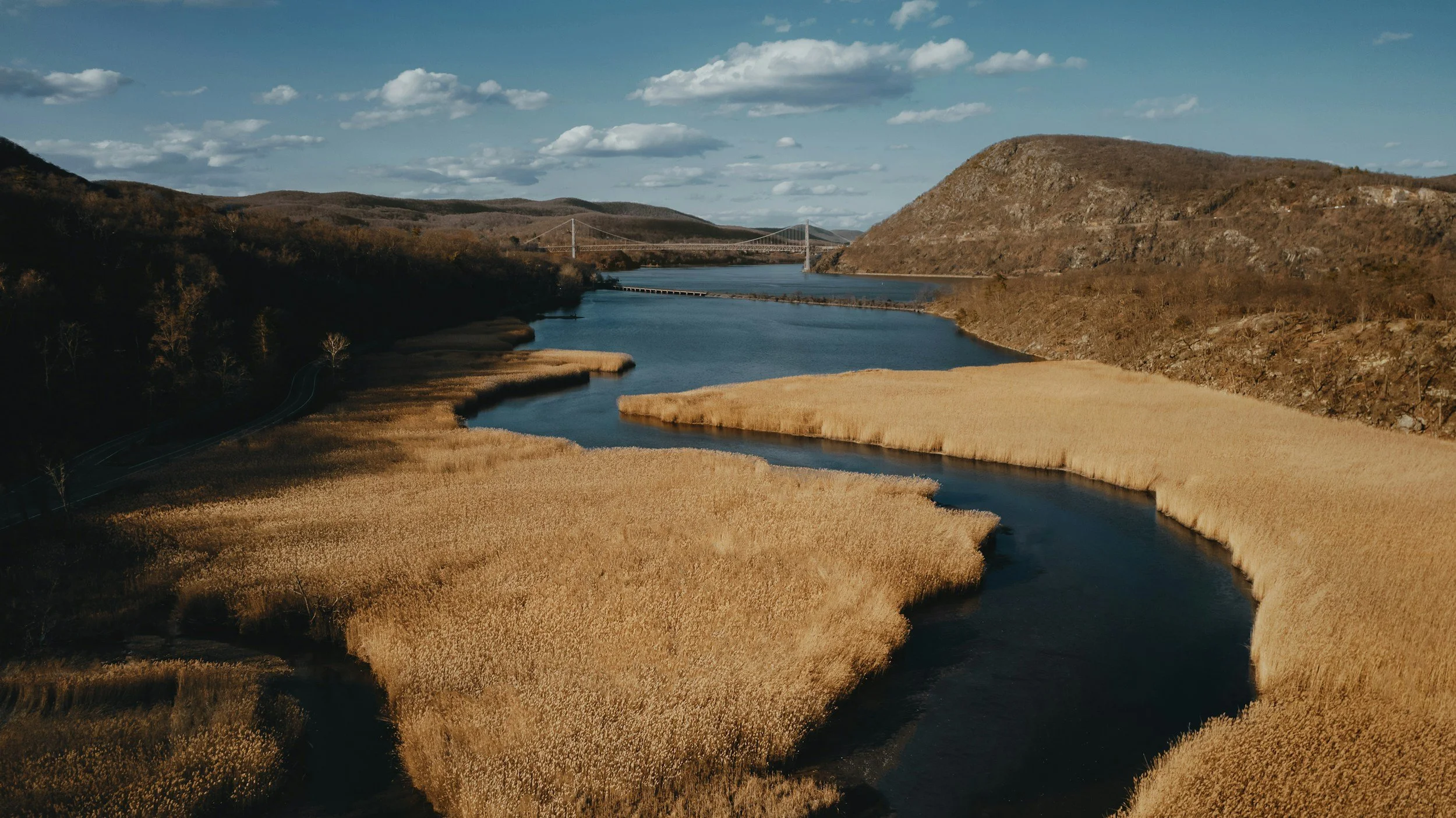 An aerial landscape photograph of the Hudson River winding past golden marshlands and rolling highlands, with the Bear Mountain Bridge visible spanning the river in the background under a blue sky.