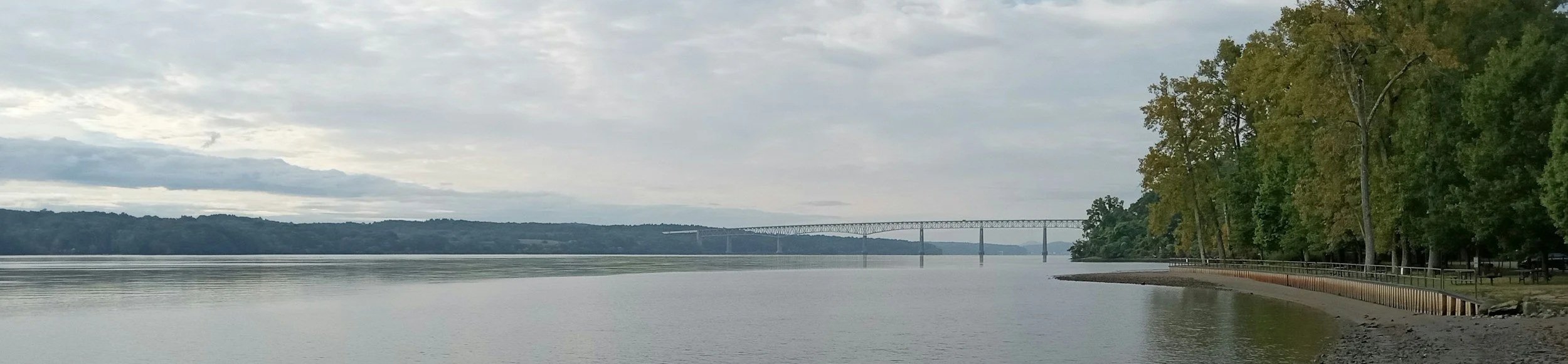 A panoramic view of the Hudson River on a cloudy day, showing the Kingston-Rhinecliff Bridge in the distance and a tree-lined shore in the foreground.