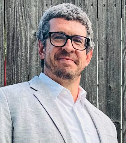 A headshot of Jake Perry, a smiling man with salt-and-pepper hair, glasses, and a light beard, wearing a gray blazer and white shirt.
