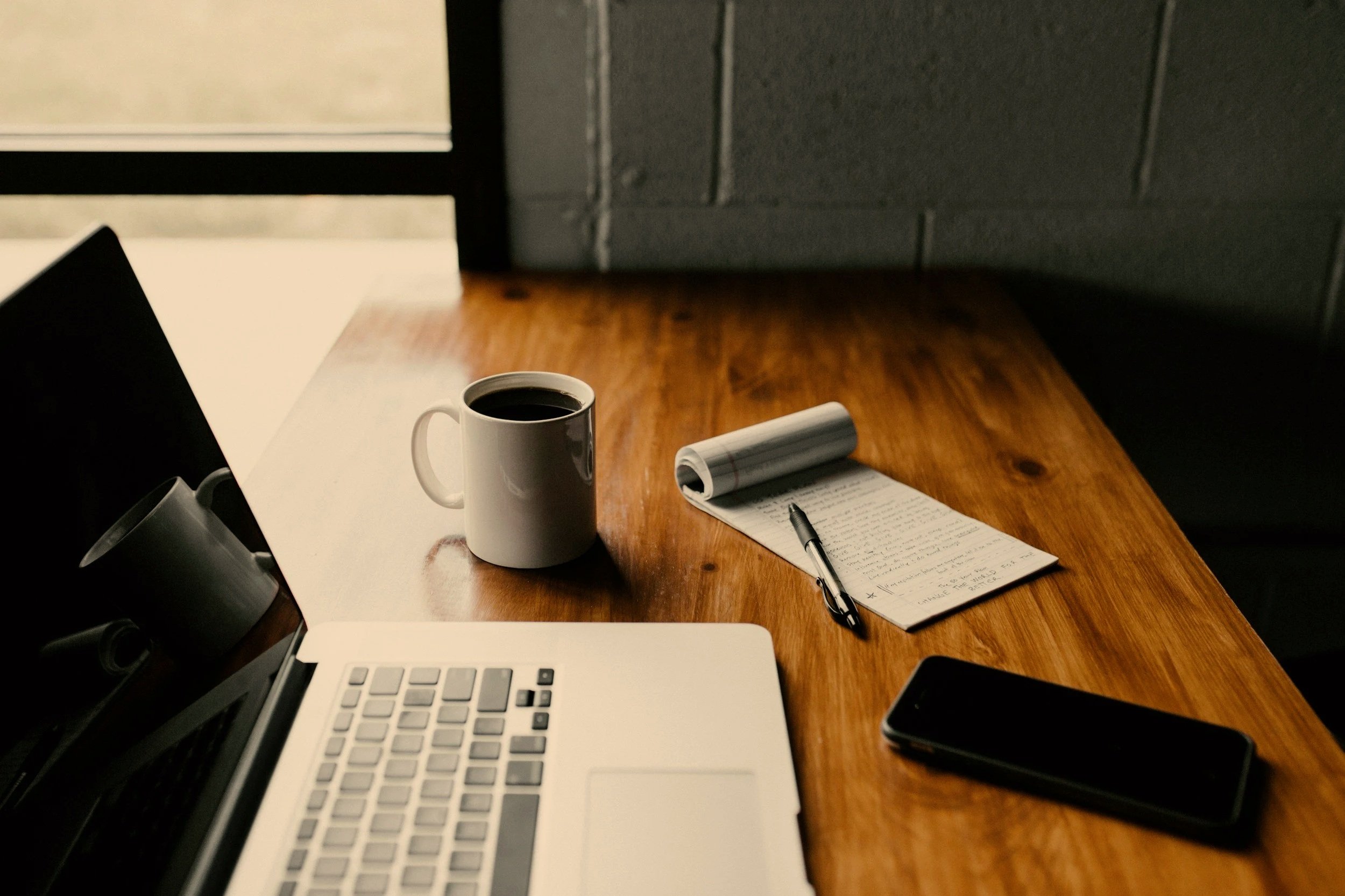 A high-angle view of a warm, wooden desktop workspace, featuring a white ceramic mug filled with black coffee, a small pen resting on a rolled open notepad with handwritten notes, and a laptop screen visible in the foreground.