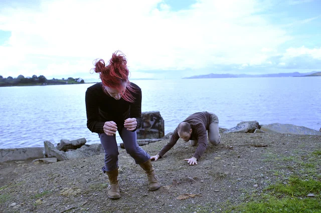 Two dancers crouched low on dirt and rock near the water of the SF Bay.