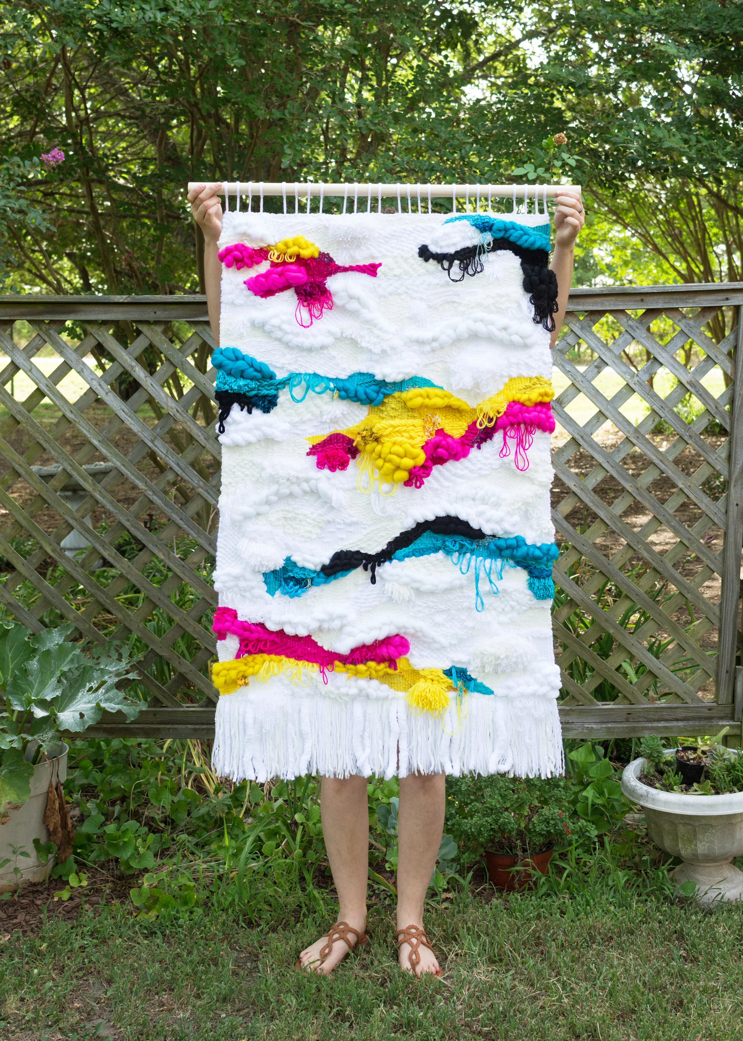 Photo of the full weaving being held up outside by the artist  in front of a garden and fence.