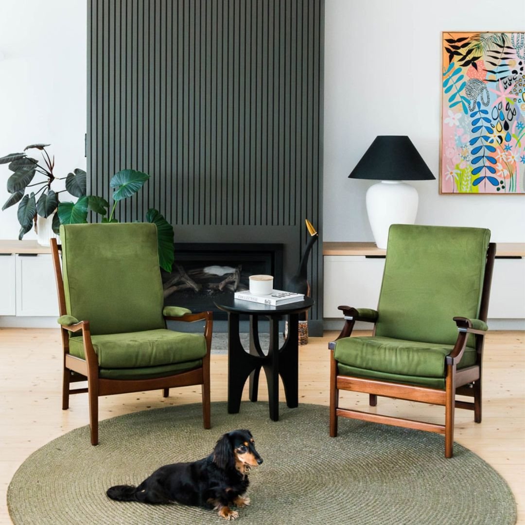 Two green velvet armchairs, one cozy pup, and a whole lot of soul 💚⁠
⁠
This space in front of the fireplace is all about balance&mdash;earthy tones, bold textures, and playful art. The slatted fireplace surround adds a modern edge, while vintage cha