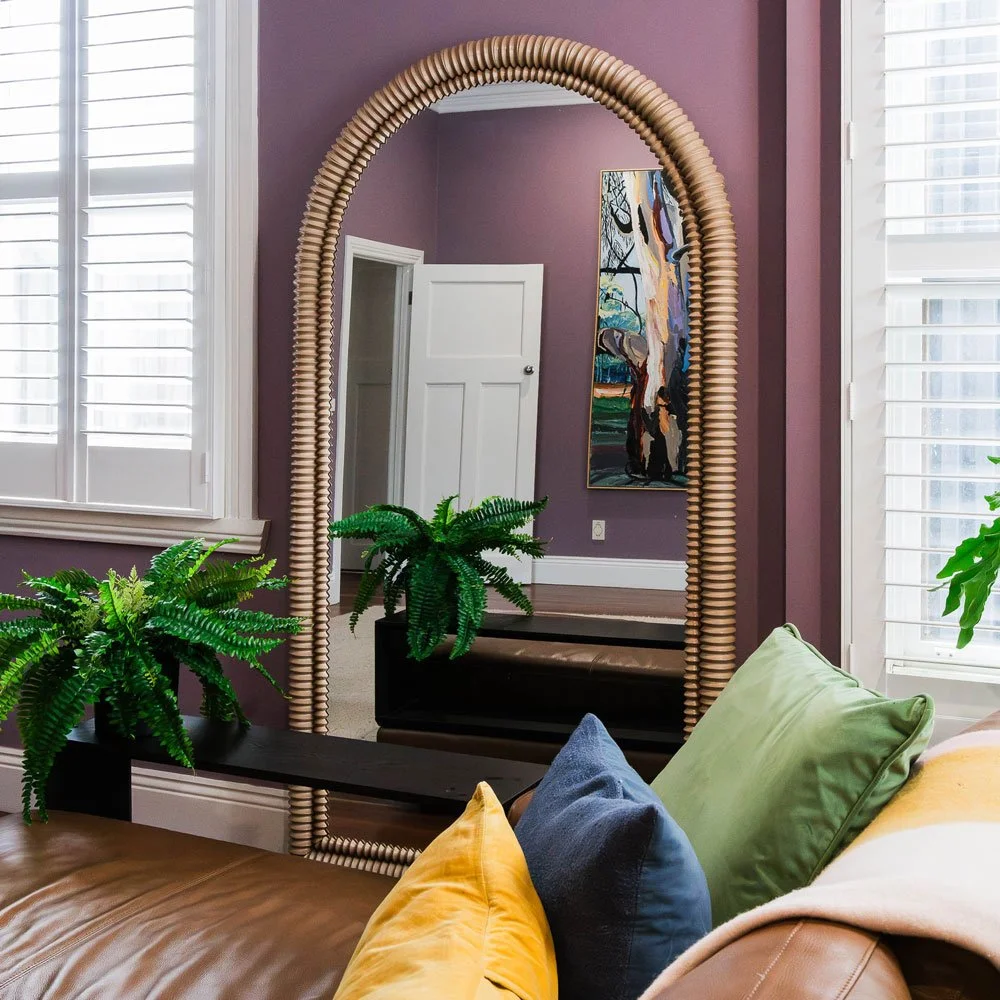 Large arched mirror reflecting art and a white door in a living room with mauve walls
