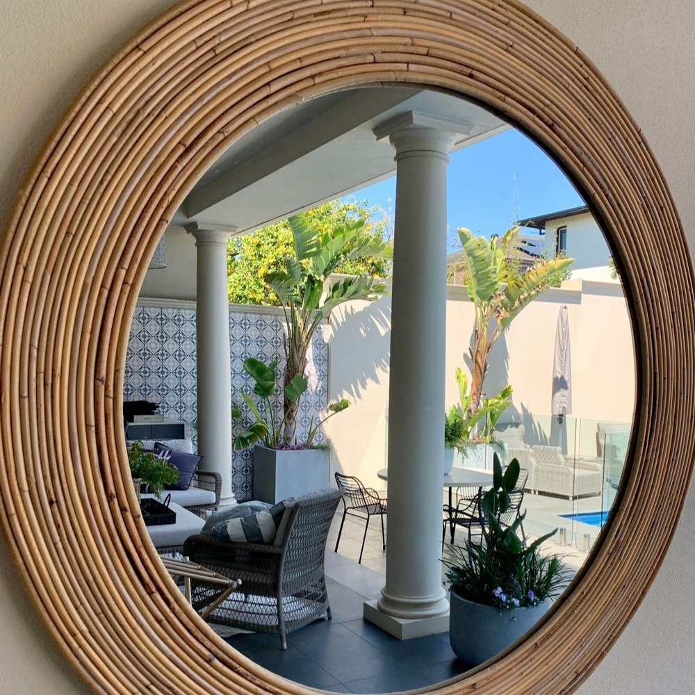 Large outdoor round rattan mirror reflecting patio furniture by a pool designed by Hue and Space Interior Design
