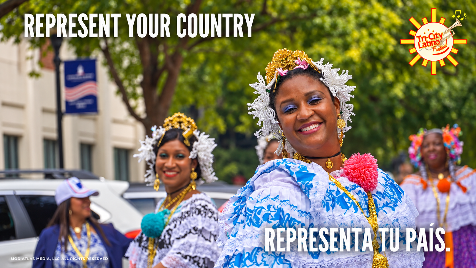 Women in traditional cultural costumes smiling and dancing during a festival, with a focus on a woman in blue and white dress adorned with gold jewelry and floral headpiece, with others dressed similarly in the background, in an outdoor urban setting.