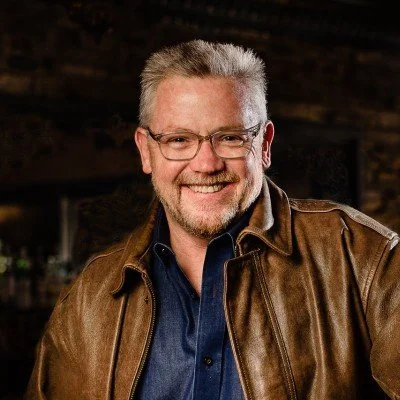 A smiling middle-aged man with gray hair and glasses, wearing a brown leather jacket over a dark blue shirt, standing indoors with a dark, blurred background.