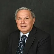 A middle-aged man with gray hair wearing a black suit, white shirt, and patterned tie posing against a dark background.