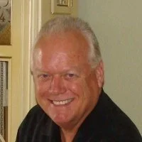A smiling middle-aged man with short gray hair wearing a dark shirt, indoors, in front of a light green wall.