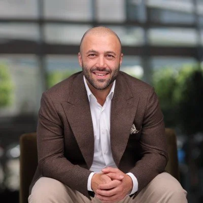 A smiling man with a beard, wearing a brown blazer and white shirt, sitting outdoors in front of a modern glass building.