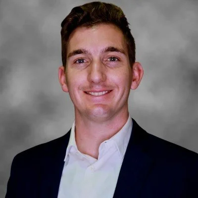 A young man with short brown hair and a big smile, wearing a white shirt and dark suit jacket, standing against a gray studio background.
