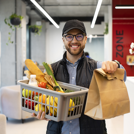 Smiling man in cap and glasses holding a grocery basket filled with fruits and vegetables, and a paper bag in a modern grocery store.