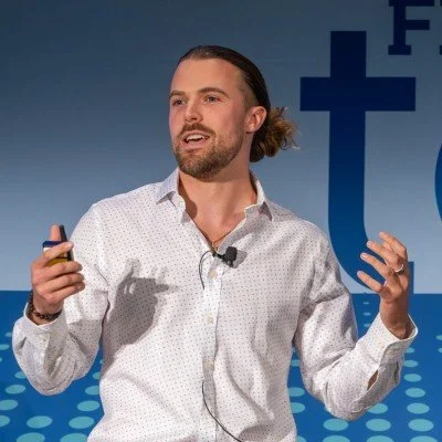 A man with long hair tied back, wearing a white shirt, speaking at a conference with a blue background featuring a large cross and the word 'Faith'.