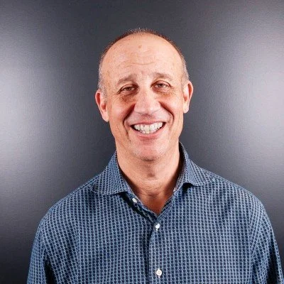 Smiling middle-aged man with short hair in a blue patterned button-up shirt against a gray background.