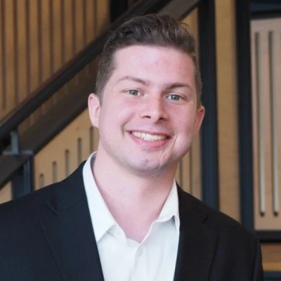 A young man with short brown hair smiling, dressed in a white shirt and dark blazer, standing indoors near a staircase with wooden railing.