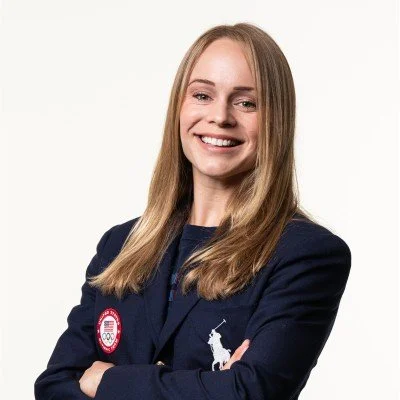Young woman with long blonde hair smiling, wearing a navy blazer with an Olympic logo and a Polo Ralph Lauren logo, standing against a white background.