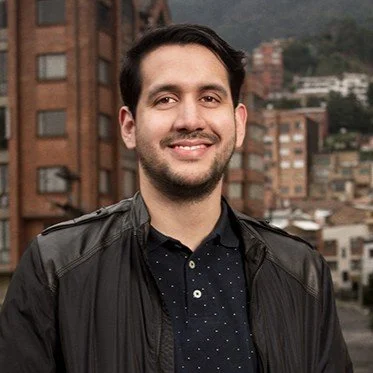 A smiling man with dark hair and a beard, wearing a black jacket and a dark shirt, standing outdoors with a cityscape and hills in the background.