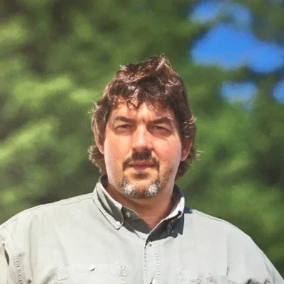 A man with dark hair and facial hair standing outdoors with trees and blue sky in the background.
