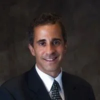 A professional man in a dark suit, white shirt, and patterned tie, smiling at the camera against a dark background.