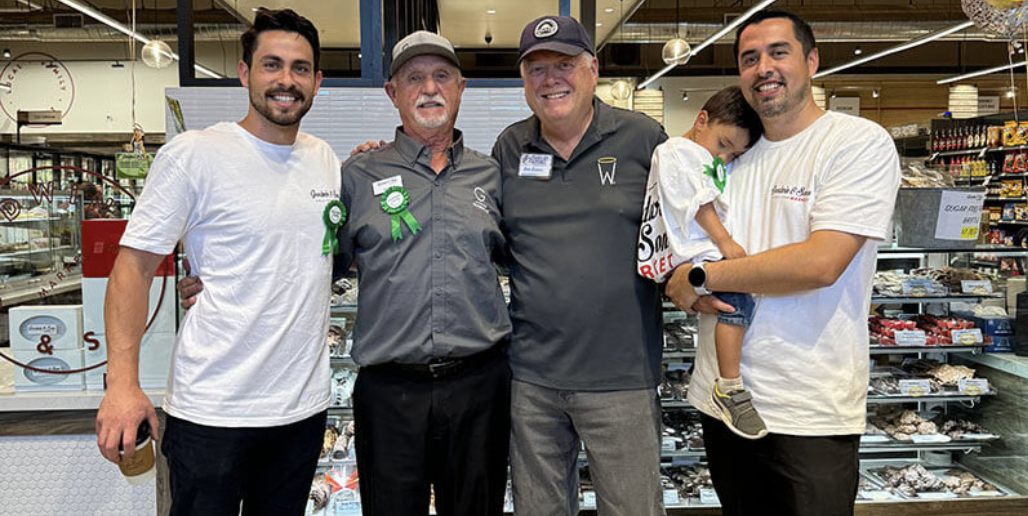 Four men and a young boy in a store, smiling for a photo. The man on the right is holding the boy. The store has shelves with various packaged items.