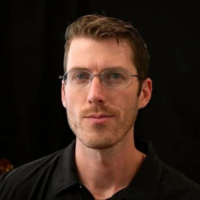 A man with short brown hair, glasses, and light facial hair wearing a black shirt against a dark background.