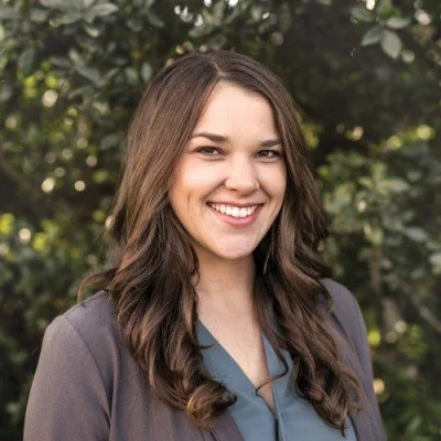 A young woman with long brown hair smiling outdoors with a background of greenery.