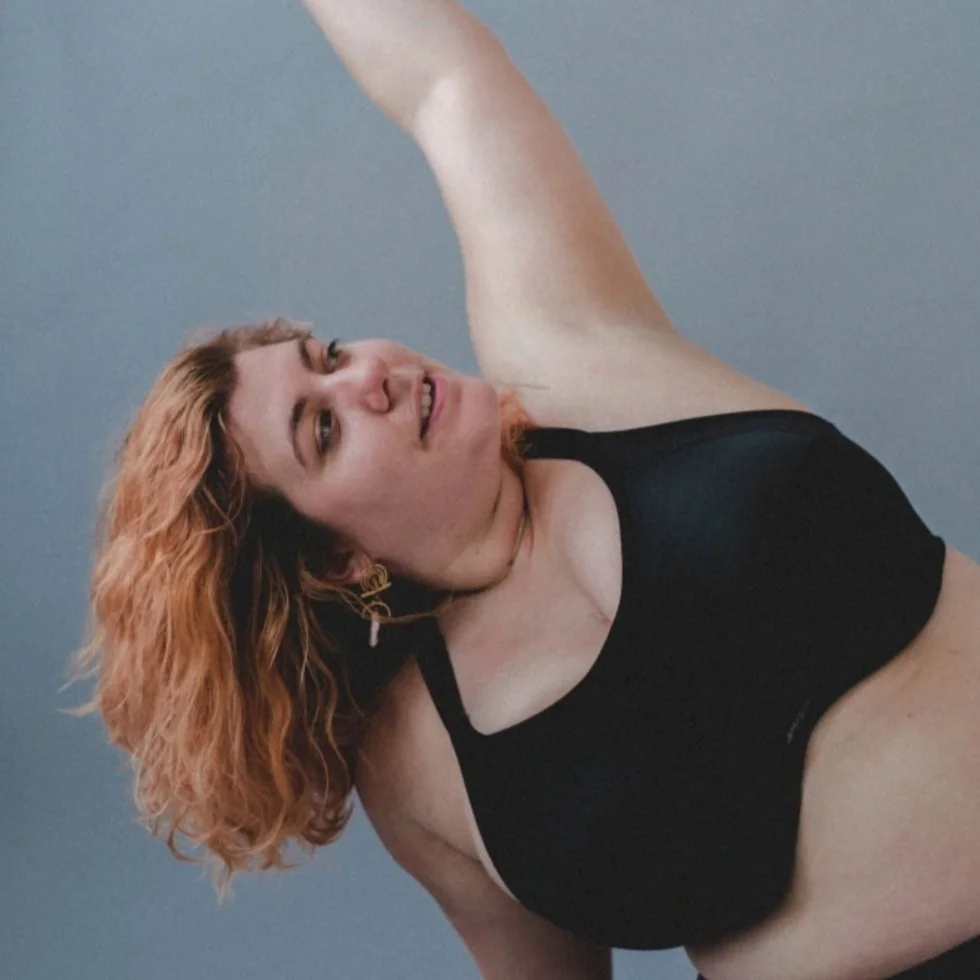A woman with curly red hair taking a selfie, wearing a black top, against a plain background.