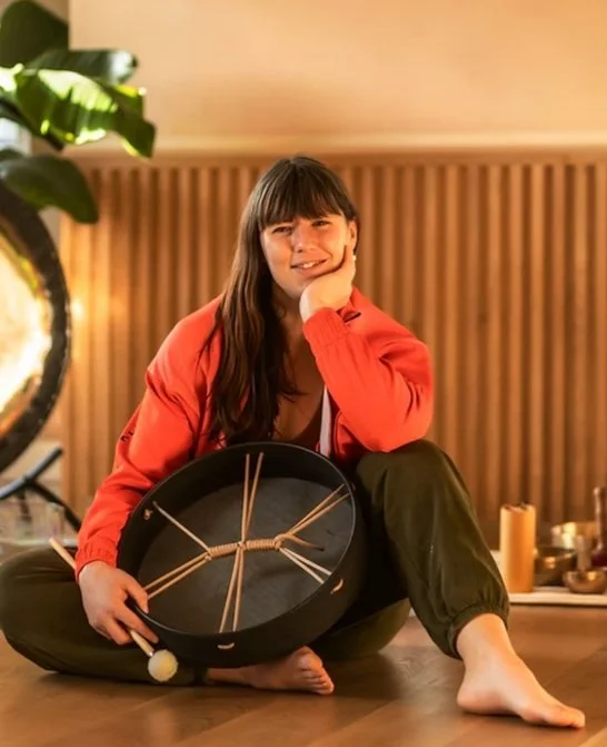 A woman sitting cross-legged on the floor, holding a circular musical instrument made of wood and string, with a relaxed and happy expression in a warmly lit room.