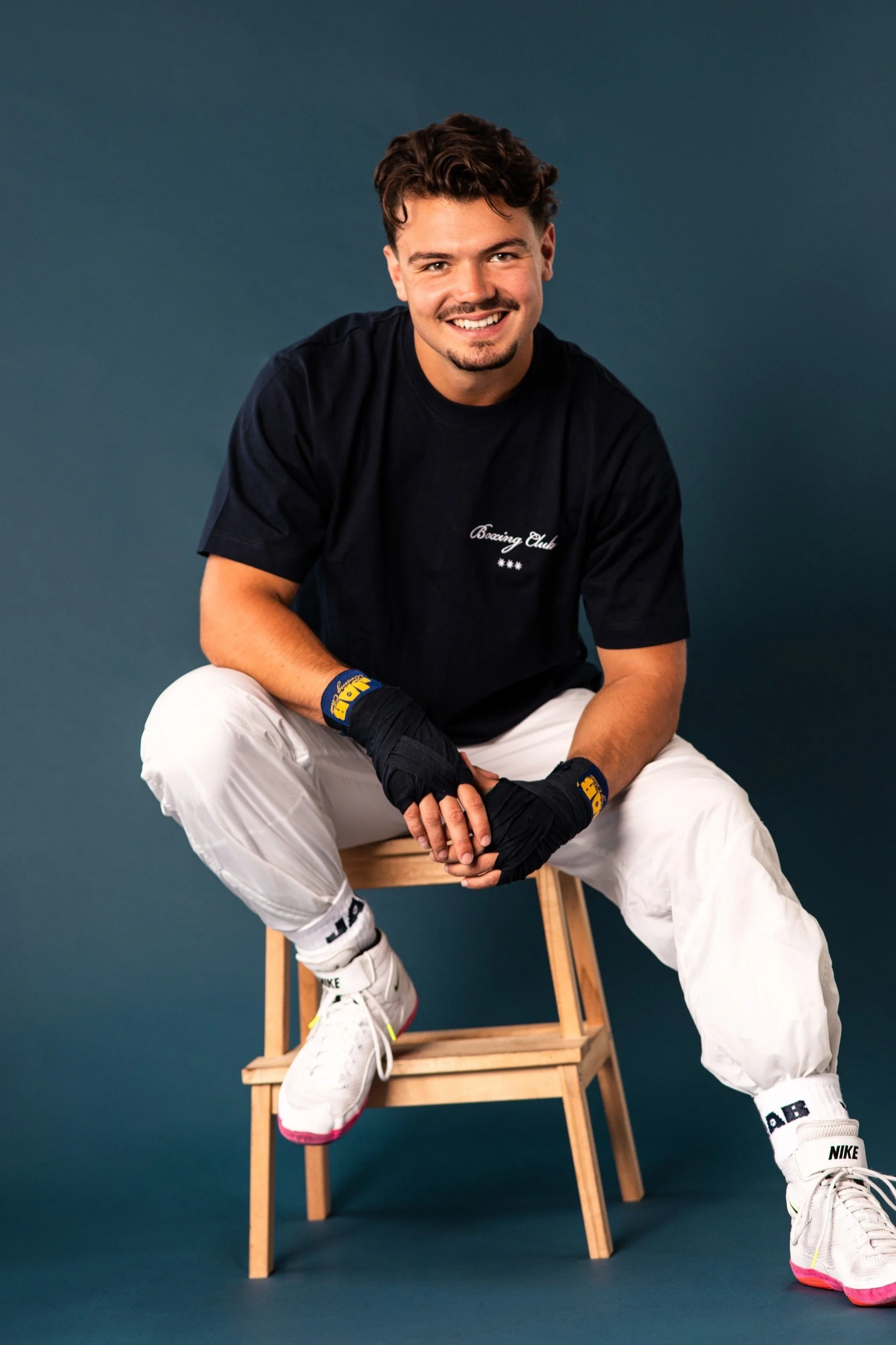 A smiling young man with curly brown hair and a goatee, wearing a navy T-shirt with 'Bacing Club' text, white pants, and white sneakers, sitting on a small wooden stool against a dark teal background.