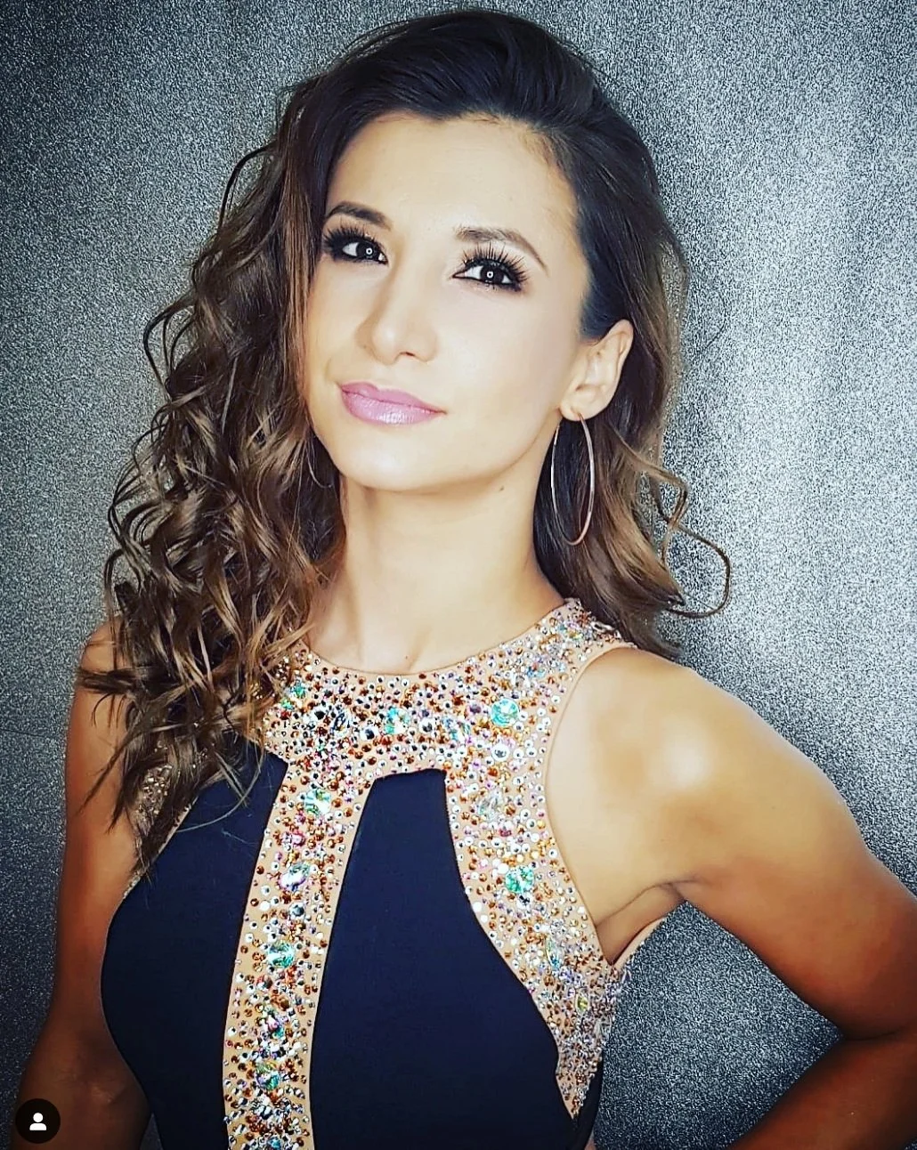 A woman with wavy brown hair, wearing a black dress with a rhinestone embellished neckline and armholes, standing against a textured gray background.